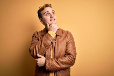 Genç, kızıl saçlı, sıradan deri ceket giyen, izole edilmiş sarı arka planı olan, eli çenesinde soru soran, düşünceli bir adam. Gülen ve düşünceli bir yüz. Şüphe kavramı.