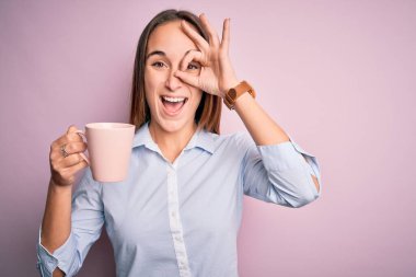 Genç ve güzel bir kadın izole edilmiş pembe arka planda dikilmiş kahve içiyor. Gülümsüyor. El ele verip parmaklarının arasından bakıyor.