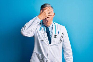 Orta yaşlı, yakışıklı, gri saçlı doktor ceketli ve mavi steteskop takmış. Şok olmuş, yüzünü ve gözlerini kapamış, utanmış bir ifadeyle parmaklarının arasından bakıyor..