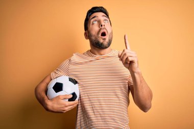 Sarı arka planda futbol topu tutan sakallı yakışıklı oyuncu hayrete düştü ve şaşırdı. Parmaklarıyla işaret etti ve kollarını kaldırdı..