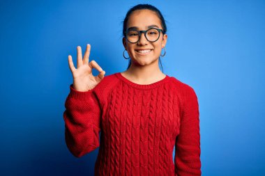 Genç, güzel Asyalı bir kadın mavi arka planda günlük süveter ve gözlük takıyor. Gülümsüyor ve el ve parmaklarıyla 