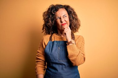 Orta yaşlı, güzel fırıncı kadın, önlük takıyor, izole edilmiş sarı arka planda duruyor, elleri çenesinde soru sorarken, düşünceli bir ifade takınıyor. Düşünceli bir yüzle gülümsüyor. Şüphe kavramı.