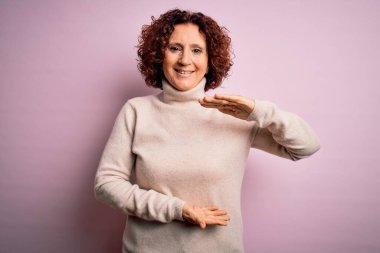 Orta yaşlı, kıvırcık saçlı güzel bir kadın pembe arka planda kazak giyiyor. Elleri büyük ve büyük beden işareti, ölçü sembolü gösteriyor. Kameraya bakıp gülümsüyor. Ölçüm konsepti.