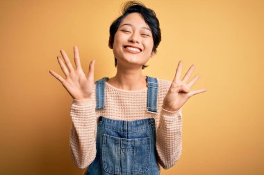 Genç, güzel Asyalı bir kız, sarı arka planda rahat bir kot pantolon giyerek dokuz numarayı gösterirken, kendinden emin ve mutlu bir şekilde gülümsüyor..