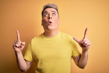 Sarıya boyanmış sarı tişörtlü genç ve modern adam şaşırmış ve şaşırmış bir şekilde yukarı bakıyor ve parmaklarıyla işaret ediyor ve kollarını kaldırıyordu..