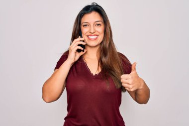 Genç İspanyol kadın akıllı telefondan konuşuyor, izole edilmiş arka planda mutlu bir şekilde gülümsüyor, işaret veriyor, parmaklarıyla başparmağını kaldırıyor, mükemmel bir işaret.