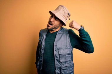 Tatilde sakallı yakışıklı turist sarı arka planda kaşif şapkası takıyor yorgun ve rahat, uykulu ve sabahın erken saatlerinde esniyor.