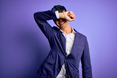 Ceket ve gözlük takan genç yakışıklı iş adamı mor arka planda gözleri kollarıyla kaplayarak neşeli ve komik gülümsüyor. Kör kavram.