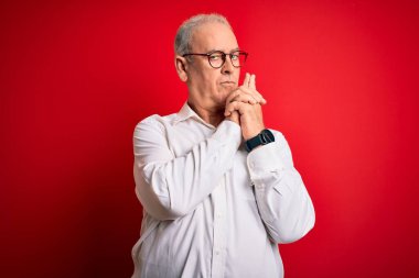 Orta yaşlı, beyaz gömlekli, kırmızı arka planda gözlüklü yakışıklı bir adam. Sembolik bir silah tutuyor. El hareketi yapıyor.
