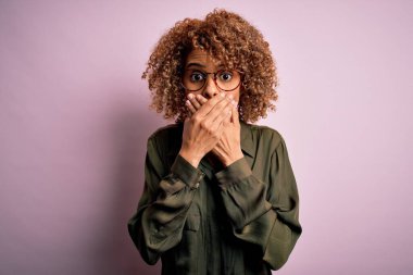 Genç, güzel, Afro-Amerikalı bir kadın pembe arka planda günlük gömlek ve gözlük takıyor. Ağzını yanlışlıkla ellerle kapatıyor. Gizli konsept.