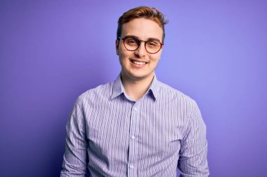 Genç, yakışıklı, kızıl saçlı, sıradan bir gömlek ve mor arka plan üzerinde gözlük takan mutlu ve havalı bir gülümsemesi olan bir adam. Şanslı kişi..