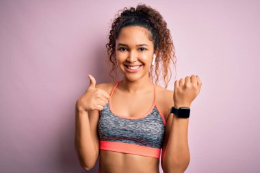 Kıvırcık saçlı güzel sporcu kadın spor giyer, spor giyer, akıllı adam kullanır, gülümser, işaret eder, parmaklarıyla başparmağını kaldırır, mükemmel bir işaret verir.
