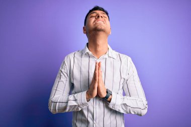 Zarif iş gömleği giyen genç yakışıklı İspanyol adam mor arka planda duruyor. Elleriyle yalvarıyor ve dua ediyor. Yüzünde umut ifadesi var. Yalvarıyorum..