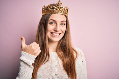 Genç, güzel, kızıl saçlı, kraliçe tacı takan, izole edilmiş pembe arka planda mutlu bir el hareketi yapan bir kadın. Kameraya bakıp başarıyı gösteren ifade onaylanıyor.