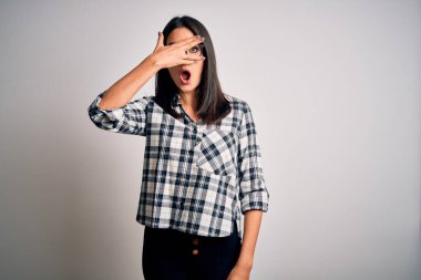 Mavi gözlü genç esmer kadın, günlük gömlek ve gözlük takıyor beyaz arka planda, şok içinde yüzünü ve gözlerini kapayarak utanmış bir ifadeyle parmaklarının arasından bakıyor..