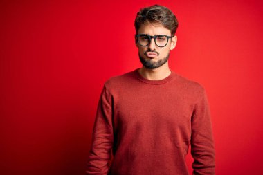 Sakallı, gözlüklü ve süveterli genç bir adam kırmızı arka planda dikilip kameraya bakarak havada öpüşüyor ve çok seksi oluyor. Aşk ifadesi.
