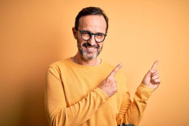 Orta yaşlı, ağarmış, günlük süveter ve gözlük takan, izole edilmiş sarı arka planda gülümseyen ve kameraya bakan, iki eliyle ve parmaklarıyla yüzünü gösteren bir adam..