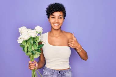 Güzel afro-amerikan kadın elinde bir buket beyaz çiçekle mor arka planda mutlu bir şekilde gülümsüyor.