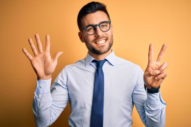 Sarı arka plan üzerinde duran kravat ve gözlük takan genç yakışıklı işadamı yedinci parmaklarını gösterip gülümserken kendinden emin ve mutlu görünüyor..