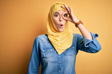 İzole edilmiş sarı arka plan üzerinde duran Müslüman başörtüsü takan genç ve güzel bir kız şaşırmış yüz ve gözlerle parmaklarından bakıyor. İnançsız ifade.