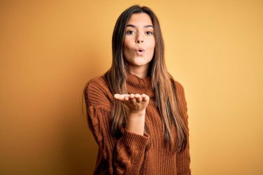 İzole edilmiş sarı arka planda dikilip el üstünde öpücükler saçan sevimli ve seksi genç ve güzel bir kız. Aşk ifadesi.