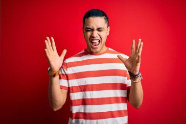 İzole edilmiş kırmızı arka planda dikilen sıradan çizgili tişört giyen genç Brezilyalı adam başarıyı kollarını kaldırmış ve kapalı gözlerle heyecanla haykırarak kutluyor. Kazanan konsept