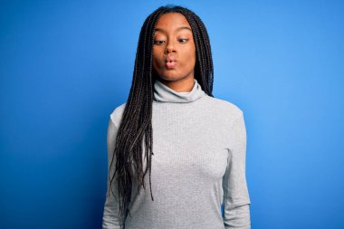 Genç Afrikalı Amerikalı kadın, mavi izole edilmiş arka plan üzerine balıkçı yaka kazak giyip dudak uçuklatıcı ve komik bir jest yapıyor. Komik ifade.