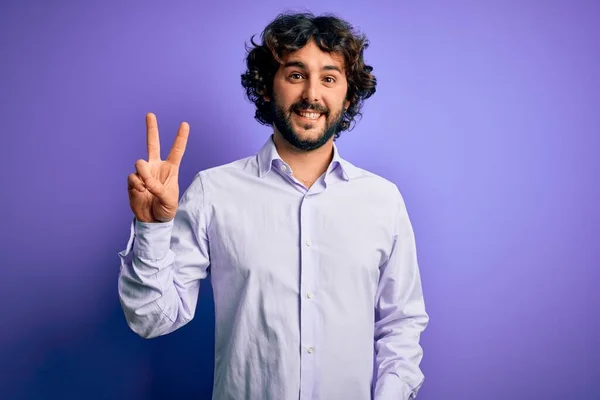 紫色の背景に立ってシャツを着てひげを生やした若いハンサムなビジネスマンは 勝利のサインをして指を示すカメラを見て笑っている 第二番 — ストック写真