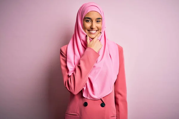 ピンクのイスラム教徒のヒジャーブとビジネスジャケットを身に着けている若い美しいブルネットの実業家は カメラが交差した腕とあごに上げ手で笑顔で自信を持って見ている 肯定的な考え — ストック写真