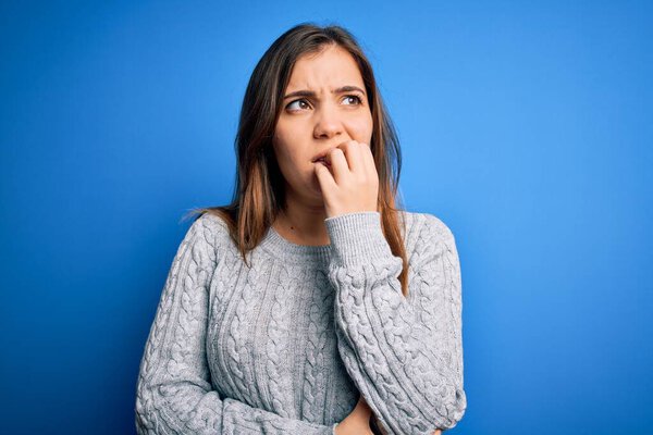 Beautiful young woman wearing casual wool sweater standing over blue isolated background looking stressed and nervous with hands on mouth biting nails. Anxiety problem.