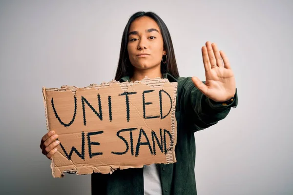 Young beautiful activist asian woman asking unity holding banner with united stand message with open hand doing stop sign with serious and confident expression, defense gesture