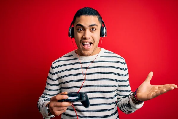 Young brazilian gamer man playing video game using joystick and headphones very happy and excited, winner expression celebrating victory screaming with big smile and raised hands