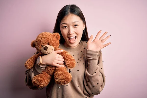 Young asian woman hugging teddy bear stuffed animal over pink isolated background very happy and excited, winner expression celebrating victory screaming with big smile and raised hands