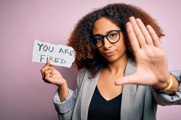 Young african american woman with afro hair holding paper with you are fired message with open hand doing stop sign with serious and confident expression, defense gesture