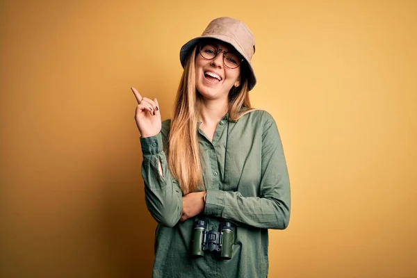 아름다운 탐험가가 모자와 안경을 미소를 쌍안경을 사용하여 손가락으로 카메라를 바라보고 — 스톡 사진