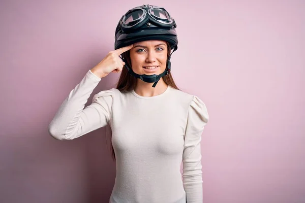 ピンクの背景の上に元ヘルメットを身に着けている青い目の若い美しいオートバイの女性1本の指で頭を指して笑顔 素晴らしいアイデアや考え 良い記憶 — ストック写真