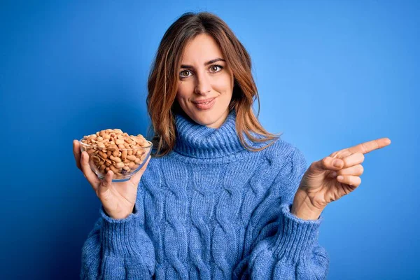 Young Beautiful Brunette Woman Holding Bowl Peanuts Blue Background Very — Stock Photo, Image