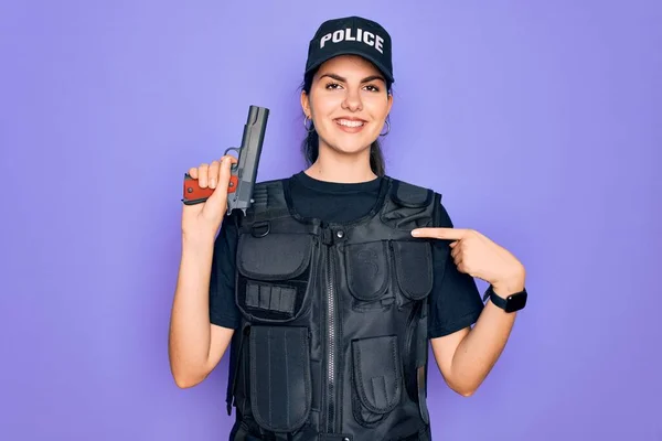 Young police woman wearing security bulletproof vest uniform and holding gun with surprise face pointing finger to himself