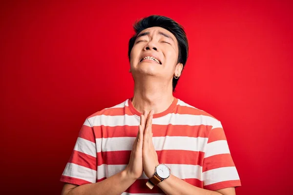 若いハンサムな中国人男性がカジュアルな縞模様のTシャツを着て 赤い背景を物乞いと手で一緒に祈って立って 顔に希望の表現は非常に感情的で心配している 物乞い — ストック写真