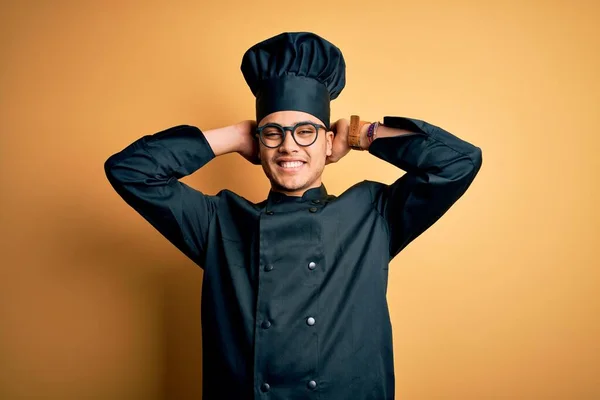 Young brazilian chef man wearing cooker uniform and hat over isolated yellow background relaxing and stretching, arms and hands behind head and neck smiling happy