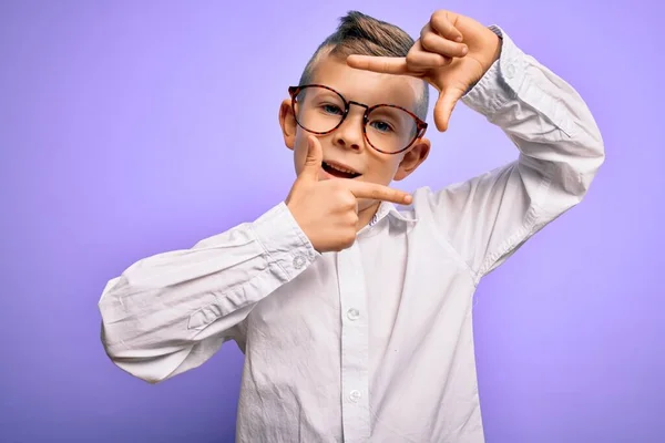 青い目をした若い白人の子供は 紫色の背景に眼鏡と白いシャツを着て 手と指で幸せな顔でフレームを作って笑っています 創造性と写真の概念 — ストック写真