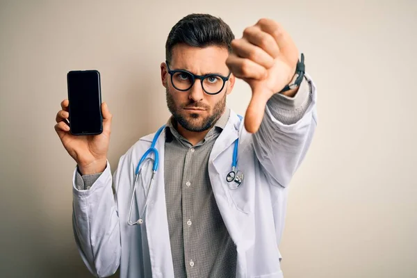 年轻的医生 戴着听诊器 在孤立的背景下展示智能手机屏幕 满脸怒容 负面迹象显示不喜欢拇指朝下 拒绝的概念 — 图库照片