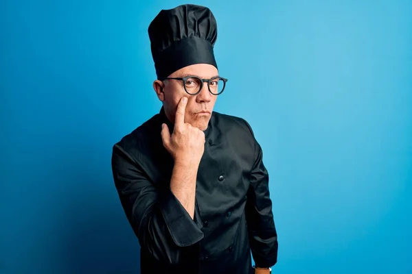 Middle age handsome grey-haired chef man wearing cooker uniform and hat Pointing to the eye watching you gesture, suspicious expression