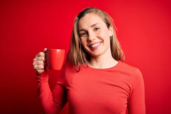 若いです美しいですブロンド女性飲むカップのコーヒースタンディング上の隔離された赤い背景で幸せな顔立ちと笑顔で自信を持って笑顔ショー歯 — ストック写真