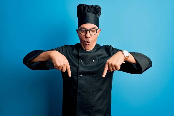 Middle age handsome grey-haired chef man wearing cooker uniform and hat Pointing down with fingers showing advertisement, surprised face and open mouth