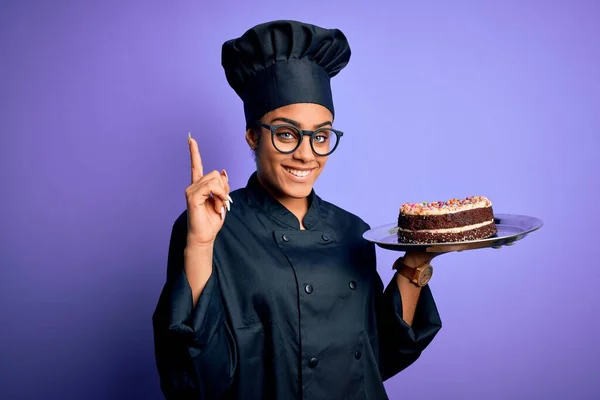 Young african american cooker girl wearing uniform and hat holding tray with cake surprised with an idea or question pointing finger with happy face, number one