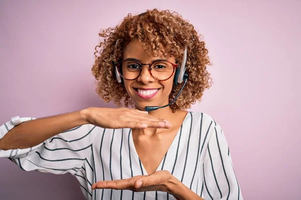 African american curly call center agent woman working using headset over pink background gesturing with hands showing big and large size sign, measure symbol. Smiling looking at the camera. Measuring concept.