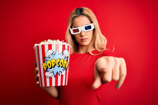 Young beautiful blonde woman watching movie using 3d glasses eating popcorns as snack pointing with finger to the camera and to you, hand sign, positive and confident gesture from the front