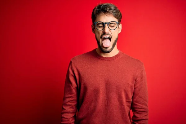 眼鏡をかけた髭を生やした若いハンサムな男と 面白い表情で幸せそうに舌を突き出している赤い背景の上に立っているセーター 感情概念 — ストック写真