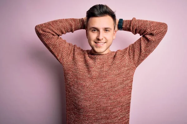 Pembe izole edilmiş arka plan üzerine sıradan bir kazak giyen, kolları ve elleri başının ve boynunun arkasında gevşeyen, gülümseyen, yakışıklı beyaz bir adam.